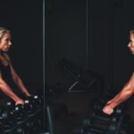 Muscle Building - woman wearing black top top holding black dumbbells standing in front of mirror